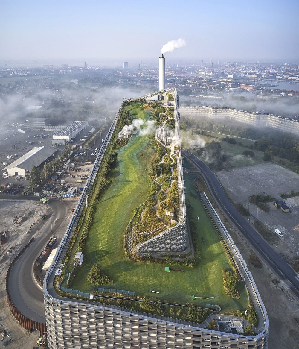 An aerial view of a large building with a green roof featuring a winding artificial ski slope and scattered vegetation; smoke emits from a tall chimney at the end. The cityscape is visible in the background.