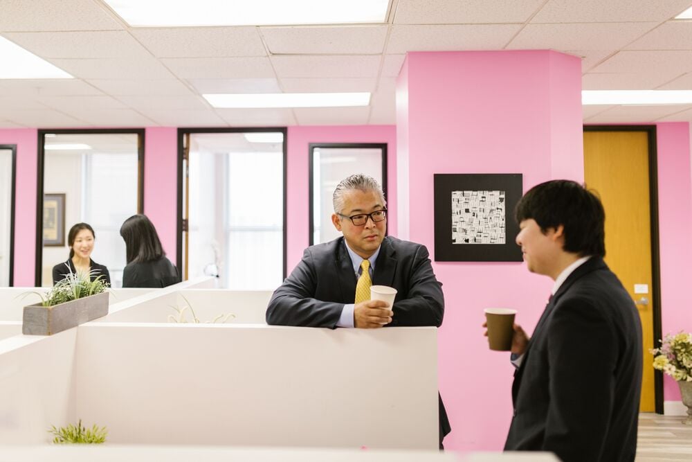 Two men in suits holding coffee cups talk in a pink-walled office. In the background, two women work at desks separated by partitions. The atmosphere is professional and bright.