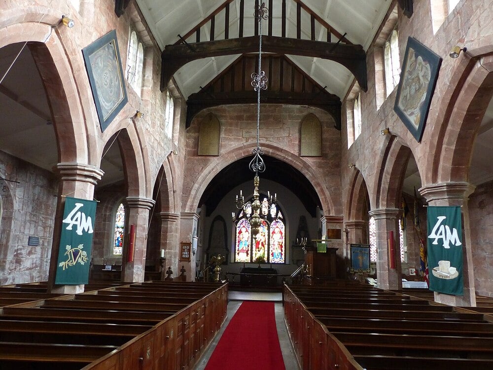 Interior of a stone church with wooden pews, red carpet aisle, arched columns, stained glass windows at the altar, chandeliers, and green banners hanging from the columns.