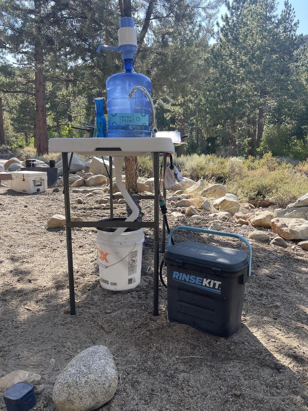 A portable camp sink setup outdoors with a water jug, soap, and bucket on a folding table, next to a black RinseKit container, surrounded by rocks and trees.