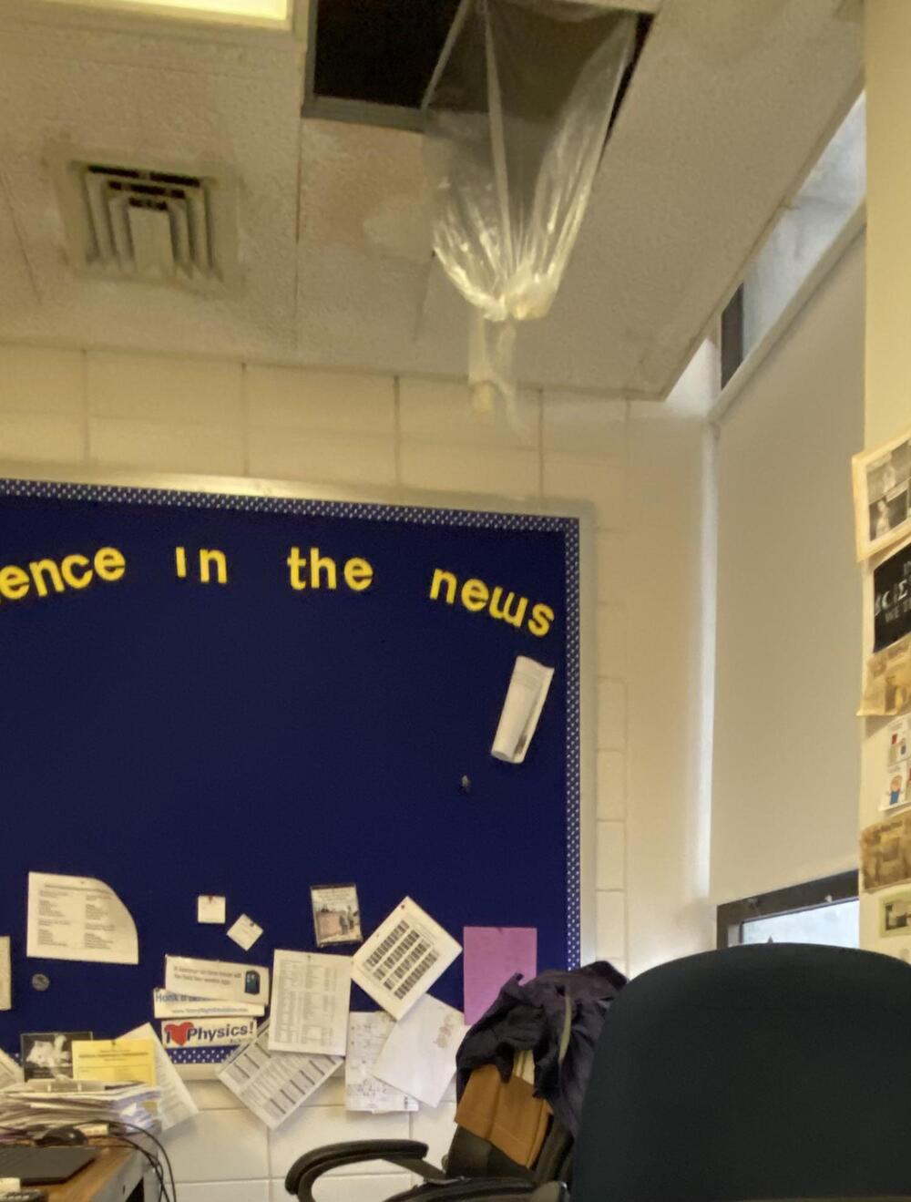 A ceiling tile is missing with plastic hanging down, possibly to catch leaks. Behind, a blue bulletin board with papers and “in the news” is visible. Part of an office chair and desk clutter are in the foreground.