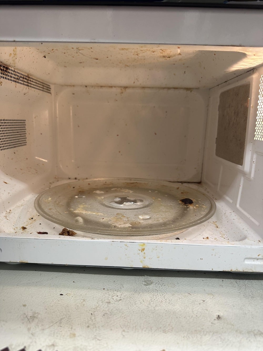 A dirty microwave interior with splatters and food stains on the walls, door, and glass turntable.