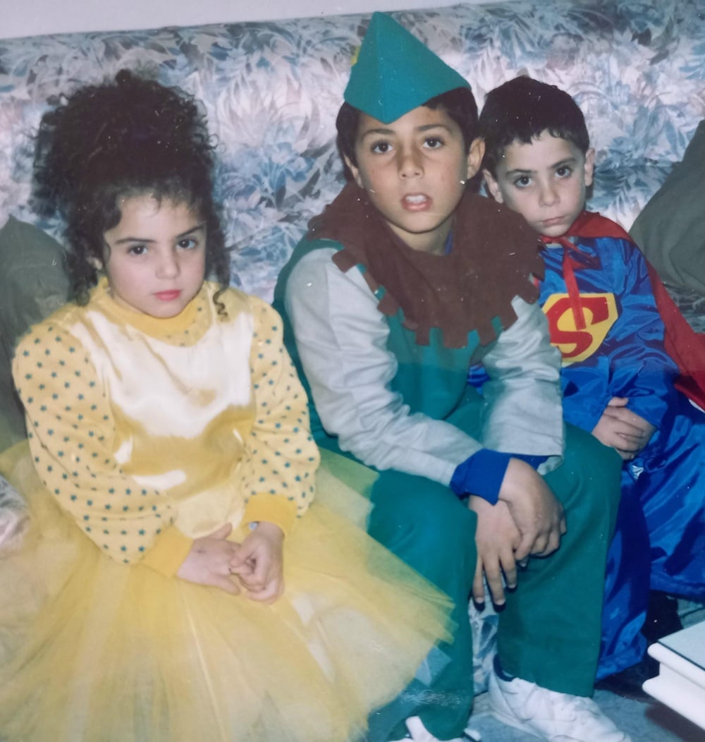 Three children sit on a floral couch in costumes: a girl in a yellow tutu, a boy dressed as Peter Pan, and another boy in a Superman outfit. They look at the camera with neutral to serious expressions.