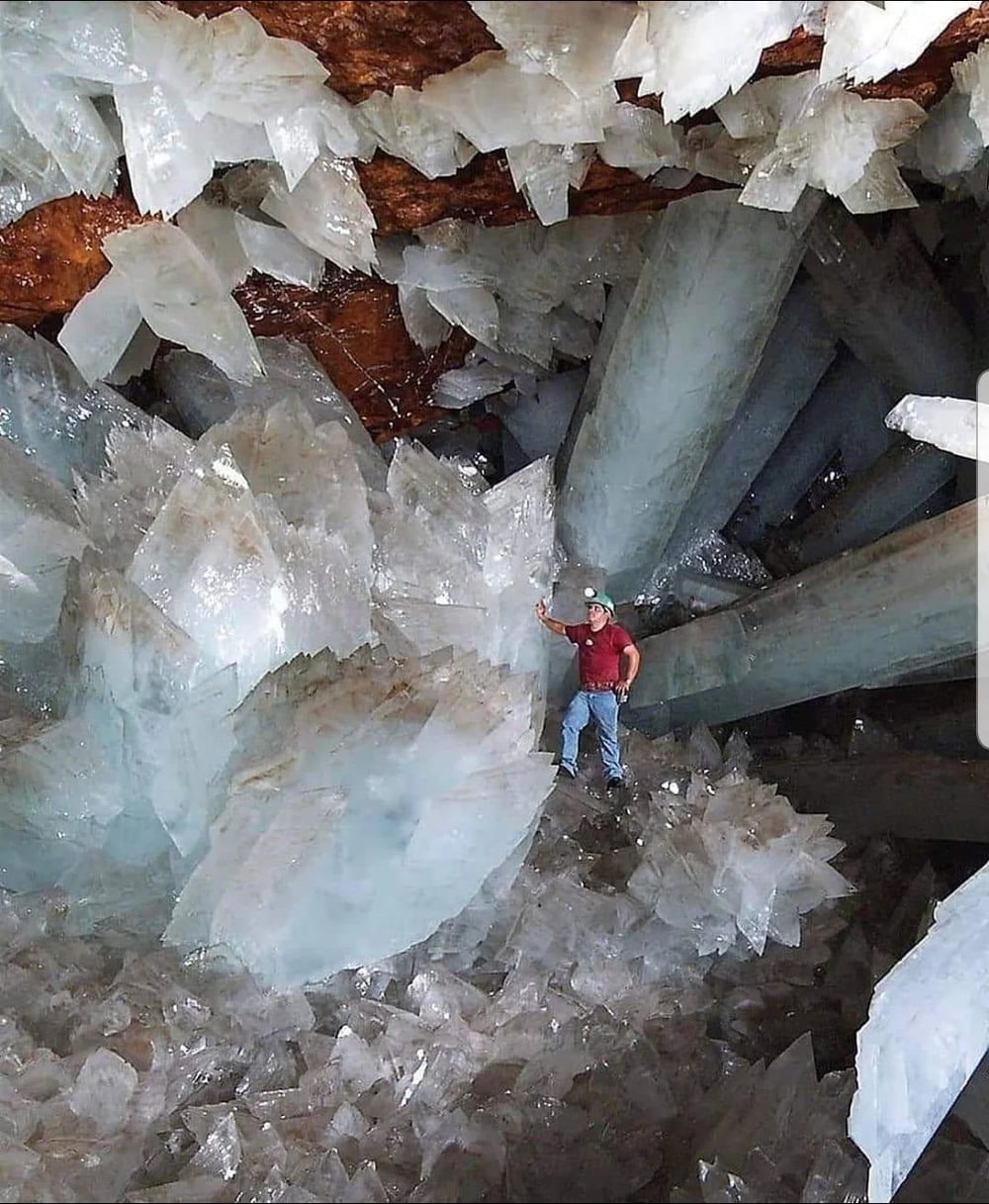 A person in a helmet stands among giant, clear gypsum crystals inside a cave. The enormous crystals surround them, highlighting the cave’s impressive and otherworldly geology.