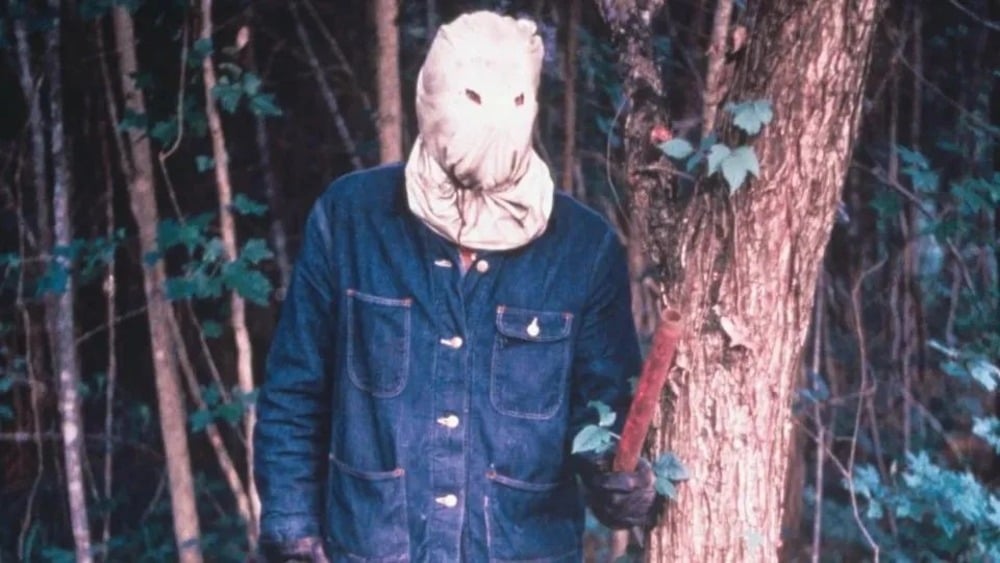 A person wearing a sack mask with eye holes, denim jacket, and holding a stick stands in a wooded area next to a tree.