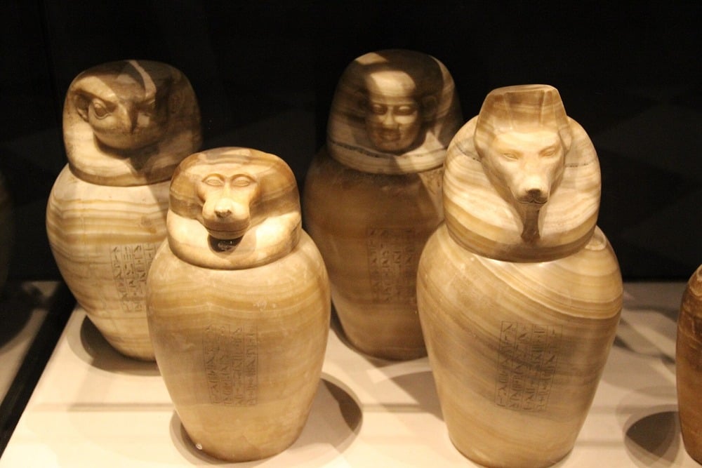 Four ancient Egyptian canopic jars made of stone, each with a different animal or human head, used for storing mummified organs. The jars are engraved with hieroglyphs and displayed in a museum setting.