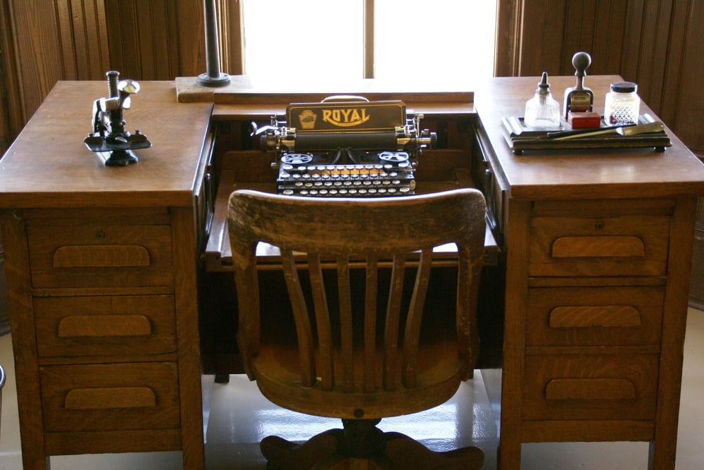 A vintage wooden desk with drawers, holding a Royal typewriter, an old-fashioned telephone, bottles of ink, a notepad, and a wooden chair, all set in front of a window.