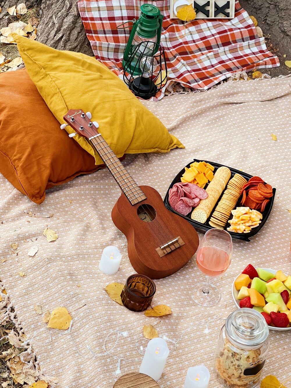 Picnic setup with pillows, a ukulele, lantern, checkered cloth, charcuterie tray, glass of pink drink, fruit bowl, jar of cookies, and candles on a beige blanket, surrounded by fallen leaves outdoors.