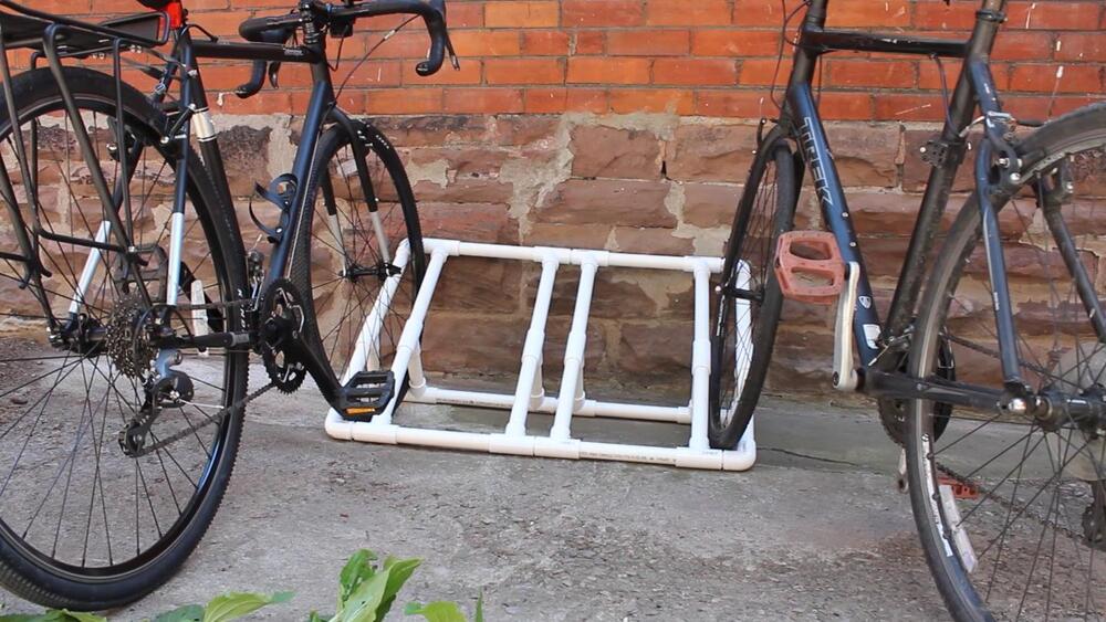 Two bicycles are parked next to a homemade bike rack made from white PVC pipes, set against a brick and stone wall on a concrete surface.