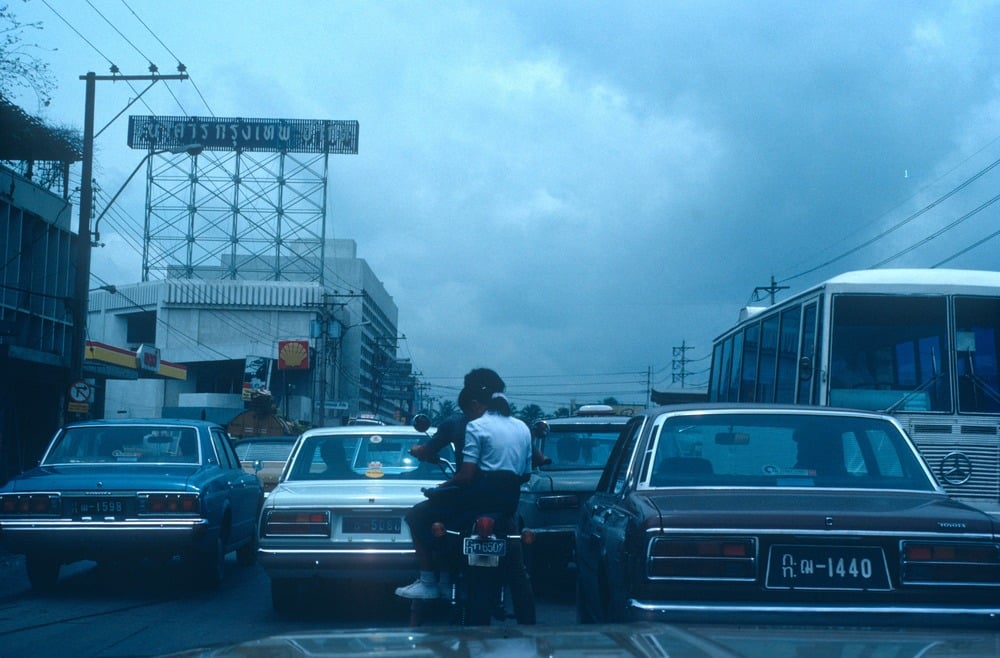 A busy city street with cars and a motorcycle in traffic, a bus on the right, and buildings with signs in Thai language under a cloudy blue sky.