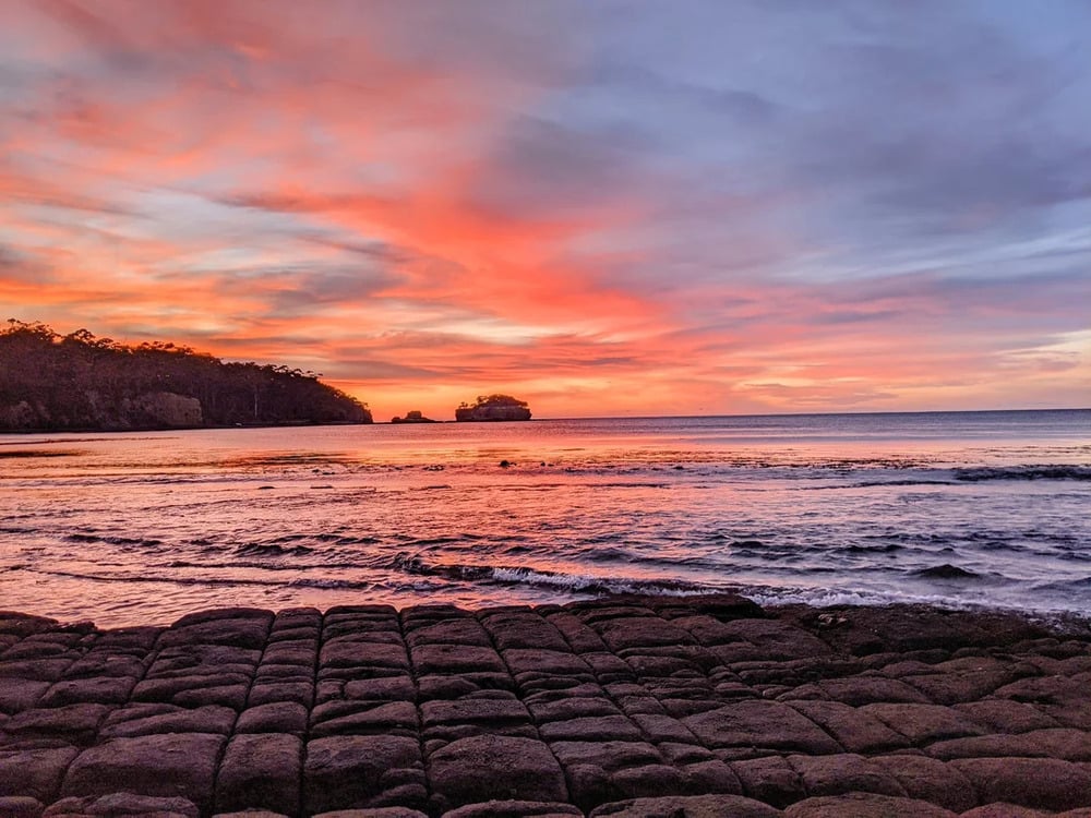 Rocky shoreline at sunset with vibrant orange, pink, and purple clouds reflecting on the calm ocean water. Small islands and silhouetted trees are visible in the distance.