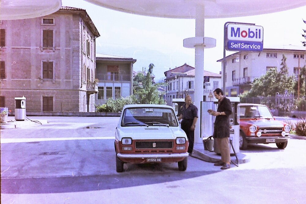 Two men stand near a white car at a Mobil self-service gas station. Another red car is parked nearby. Several buildings and trees are visible in the background. The scene appears to be from the past.