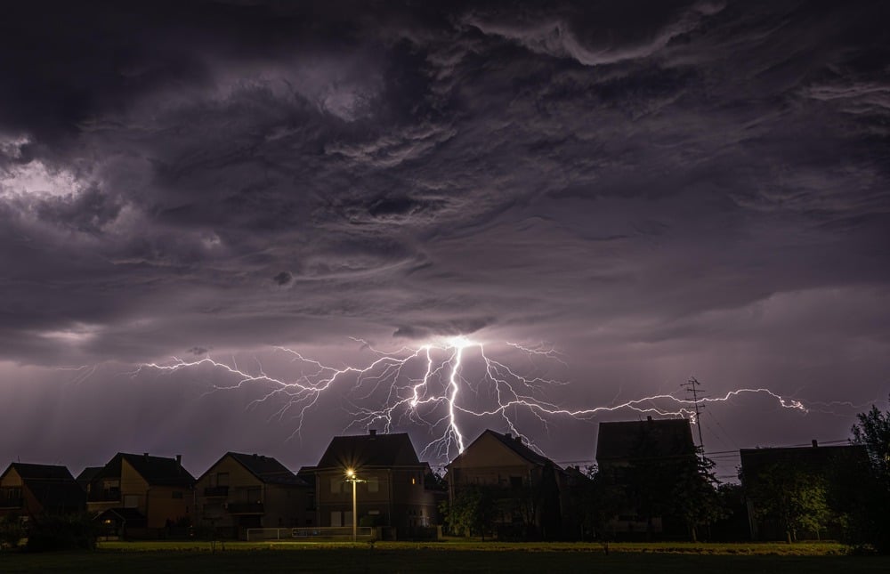 Lightning bolts illuminate storm clouds over houses at night.