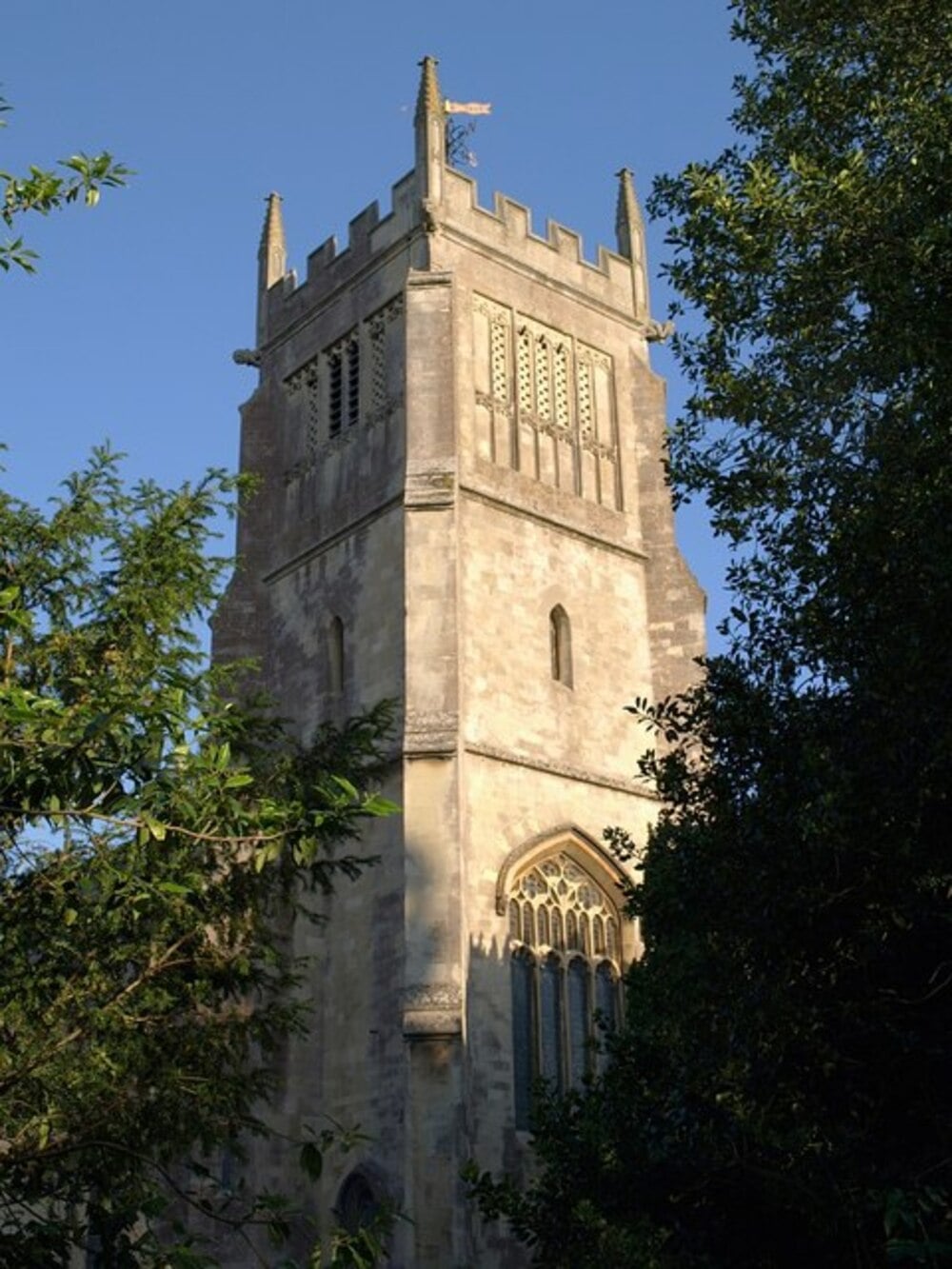 A tall stone church tower with battlements stands against a clear blue sky, partially framed by green trees.