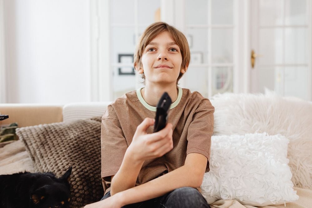 A smiling child sits on a couch holding a TV remote. A small black dog is partially visible on the left. The background features white doors and cozy pillows.