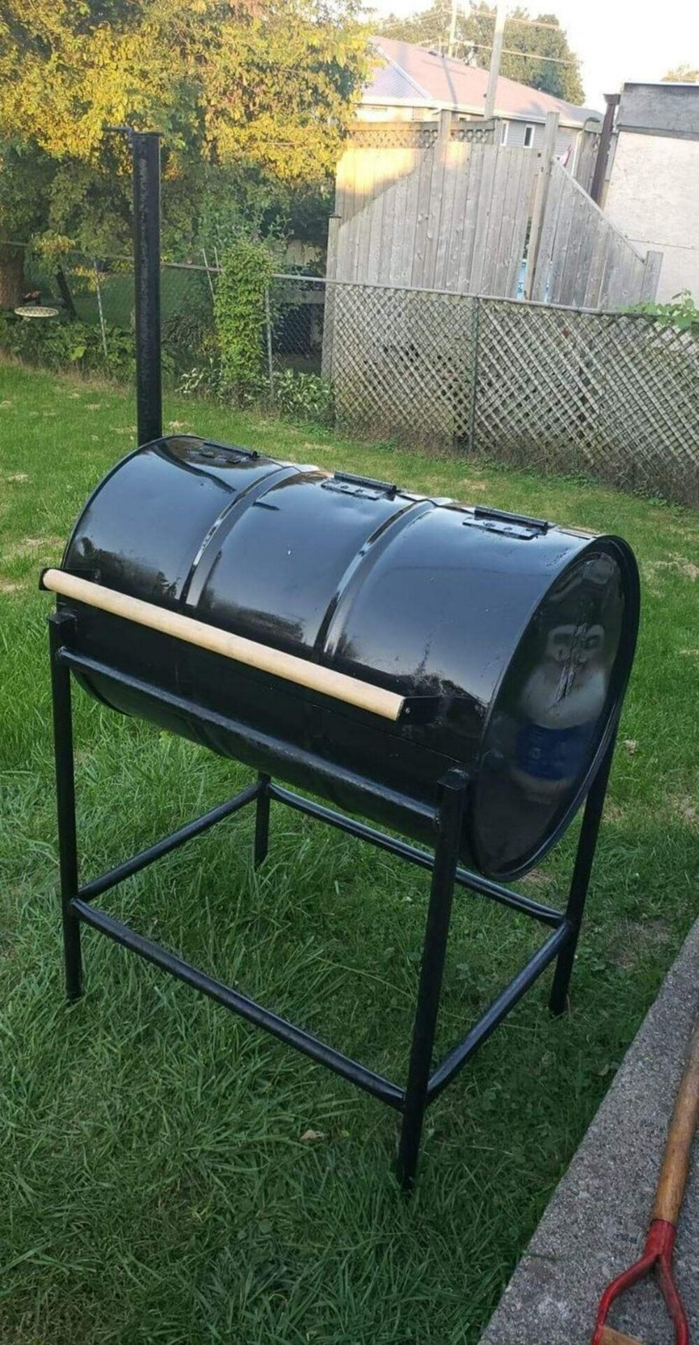 A black metal barrel mounted on a metal stand, resembling a homemade barbecue grill or smoker, sits on grass in a backyard near a fence and garden tools.