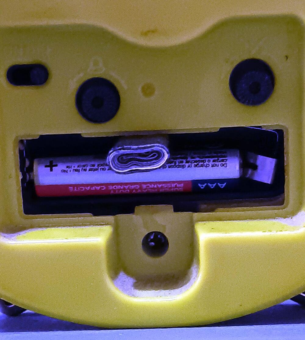 Close-up of a yellow plastic device showing a battery compartment with a single AA battery and a folded paperclip placed inside alongside the battery.