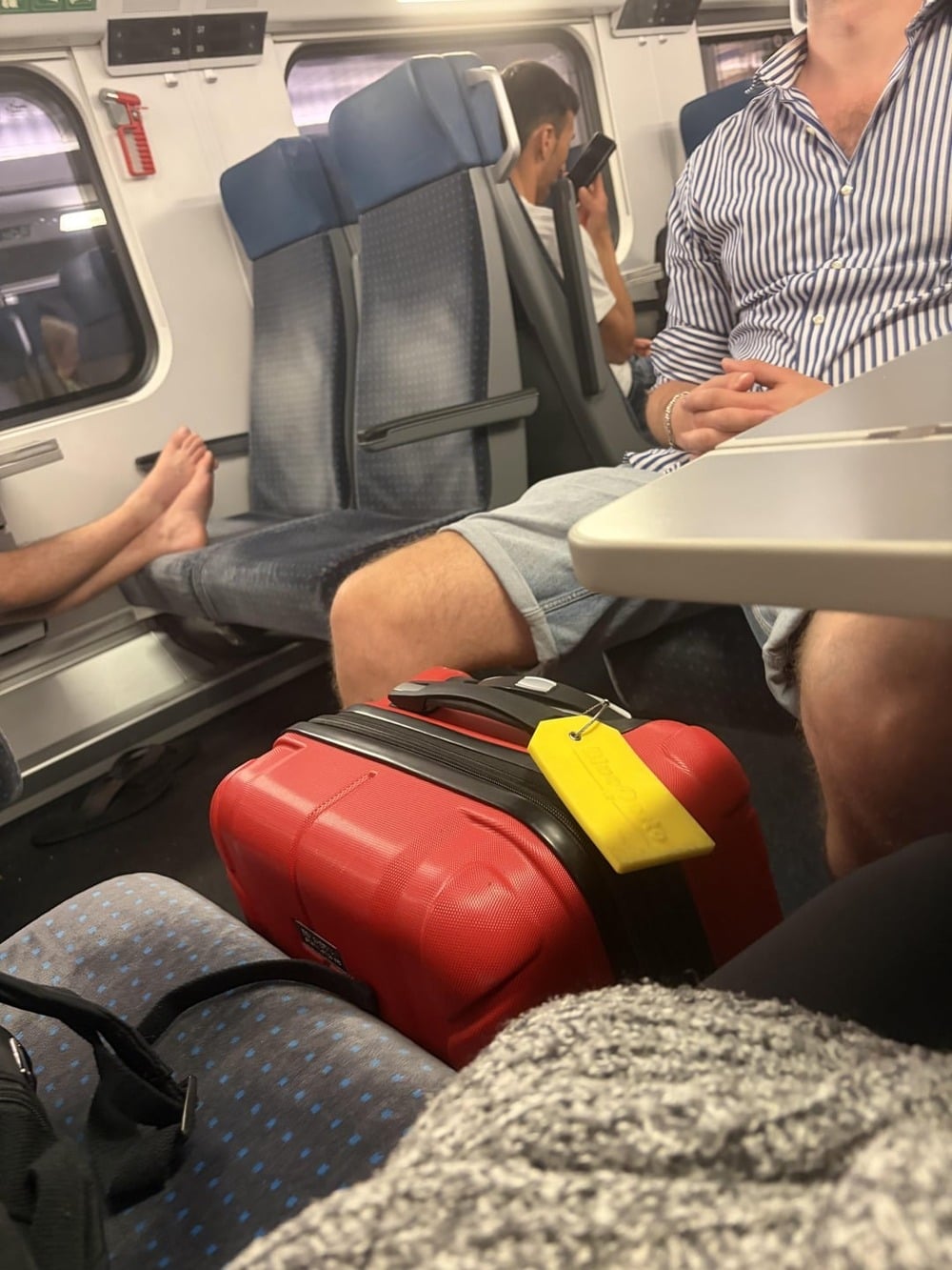 People sitting on a train; one person has feet up on a seat, another is on their phone. A bright red suitcase with a yellow tag is on the seat in the foreground. Blue cushioned train seats are visible throughout.