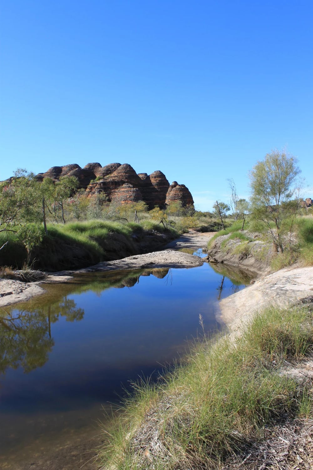 A clear stream reflects blue sky and beehive-shaped rock formations, with green shrubs and grass along the banks. The landscape is calm and sunny.