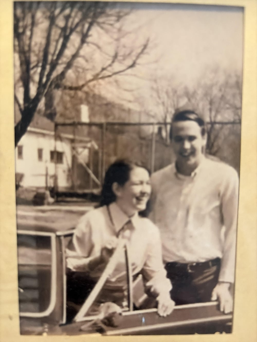 A smiling man and woman stand beside a car with trees and a building in the background. The photo appears to be old and slightly faded.
