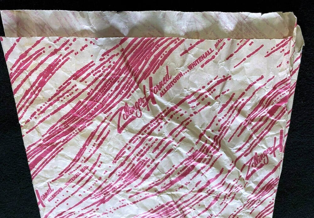 A crumpled white paper bag with red diagonal lines and the words “Zoller’s Hotel” printed repeatedly alongside city names. The bag appears empty and is set against a black background.