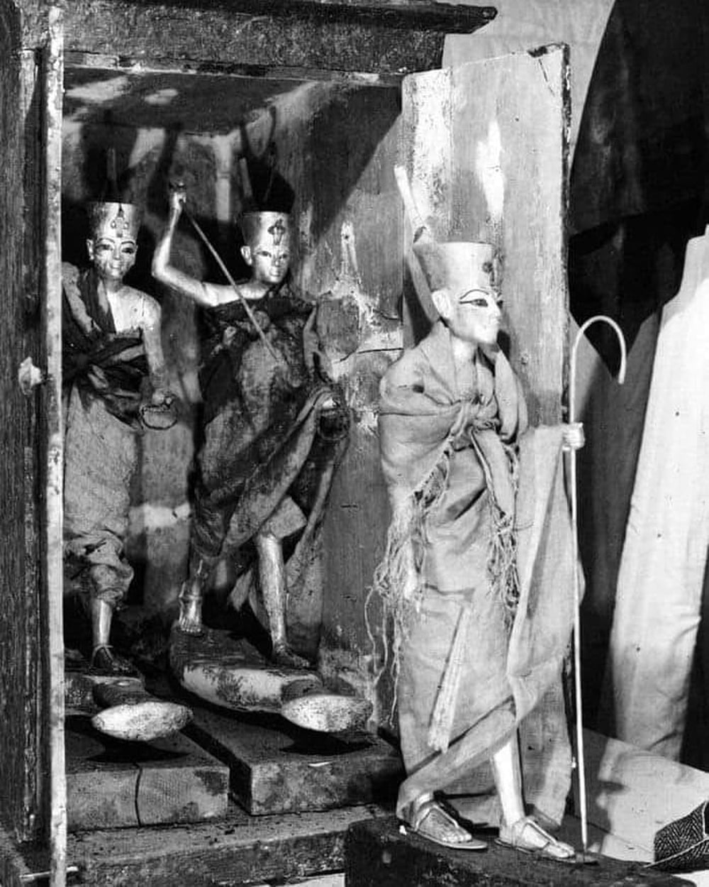 Three ancient wooden figurines dressed in robes and headdresses stand inside a box-like structure, with two holding staffs and one holding a stick, appearing to walk forward. The scene is in black and white.