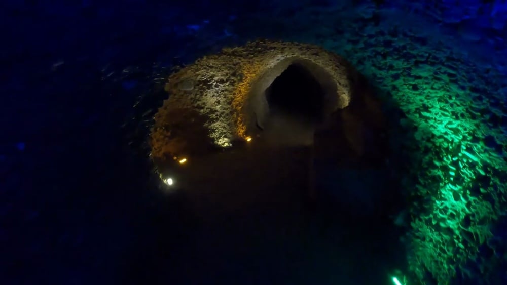 A dimly lit cave tunnel with textured walls, illuminated by small yellow and green lights, leading into a dark passage.
