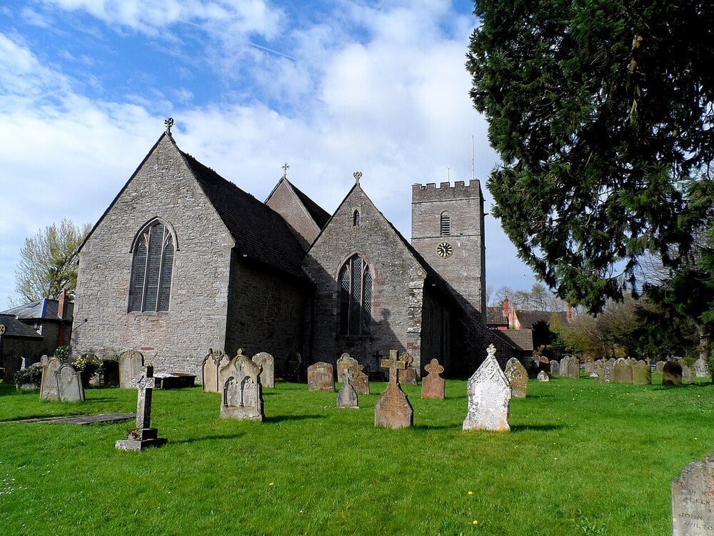 A stone church with arched windows and a square clock tower stands amid a green graveyard with old tombstones under a partly cloudy sky.