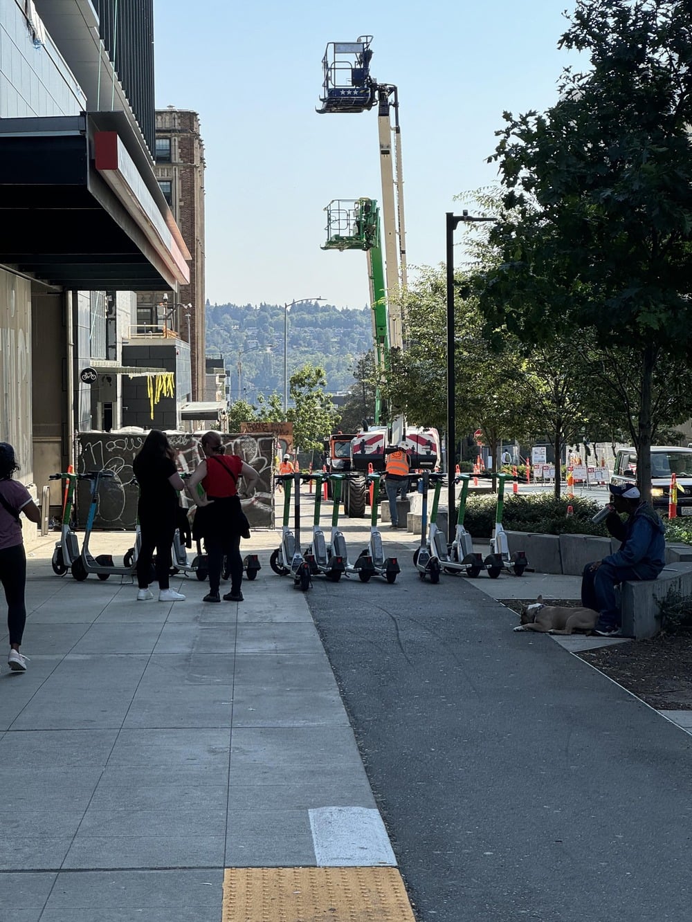 People gather near electric scooters lined up on a sidewalk, with construction equipment and workers visible in the background under a clear sky.