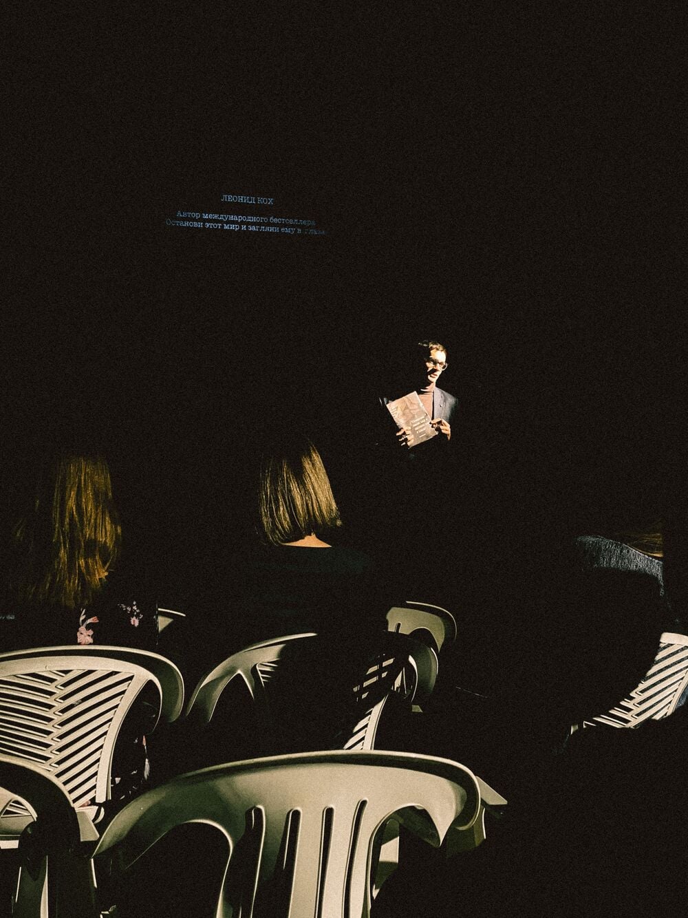 A person stands in a spotlight facing an audience seated on white plastic chairs.