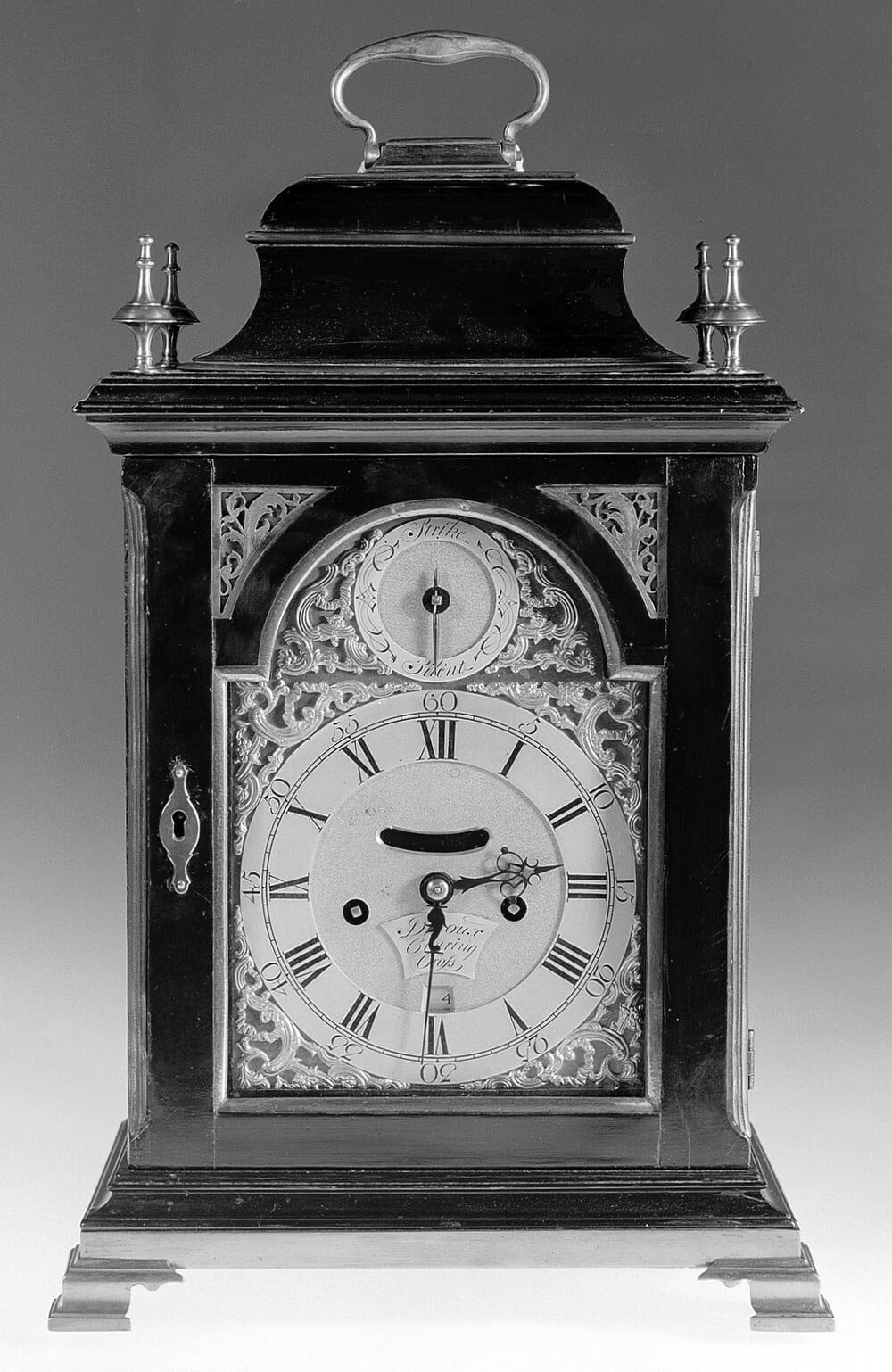 Black and white photo of an ornate antique mantel clock with Roman numerals, intricate metalwork, a handle on top, and decorative finials. The clock face shows the time as 5:43.