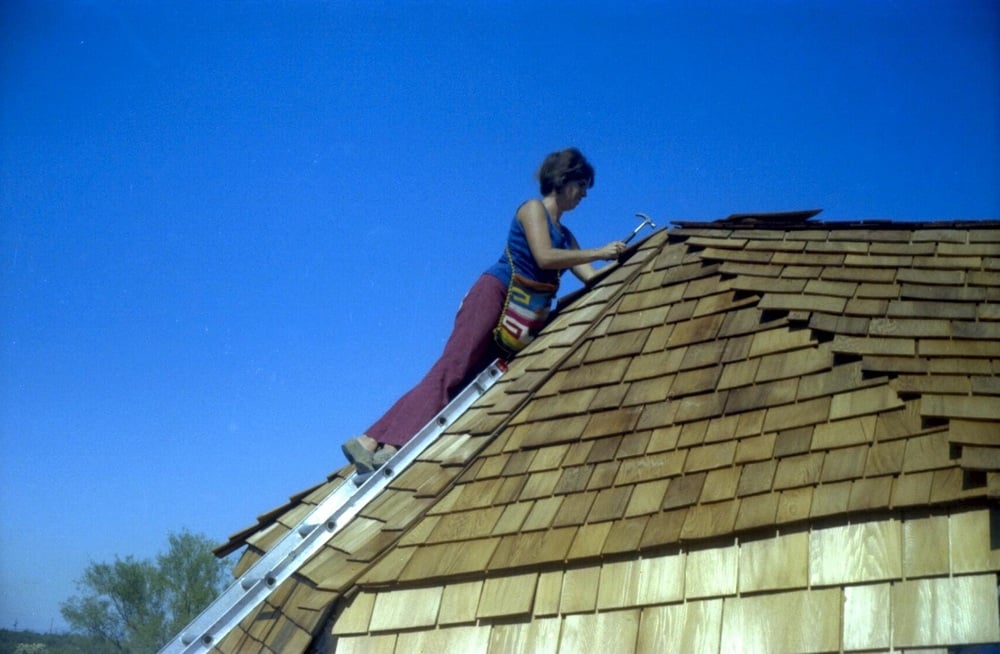 A person climbs a ladder and hammers wooden shingles onto a steep roof under a clear blue sky. They are wearing a colorful bag and red pants.