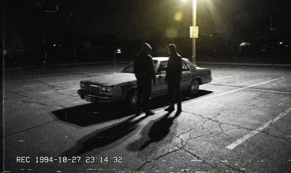 Two people in hoodies stand by a car under a streetlight in an empty parking lot at night. The image has a grainy, black-and-white, vintage camcorder aesthetic with a timestamp in the corner.