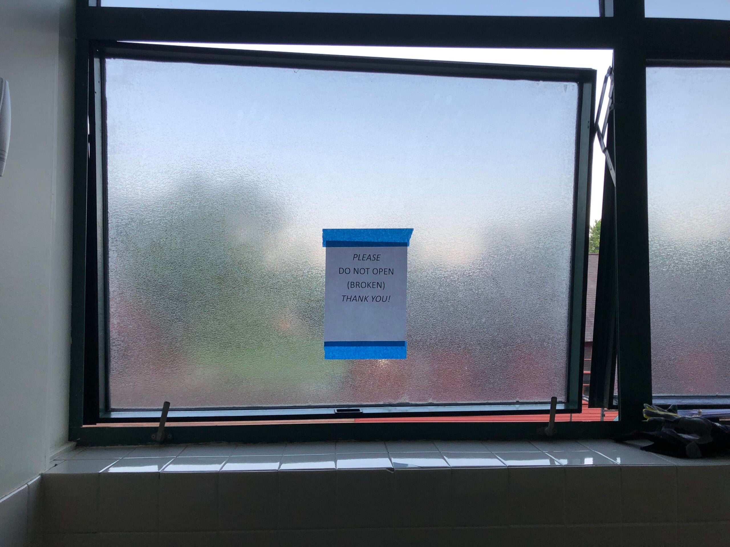 A frosted glass window, partly open, has a sign taped to it saying, “PLEASE DO NOT OPEN (BROKEN) THANK YOU!” The window is above a tiled surface, and greenery is visible outside.