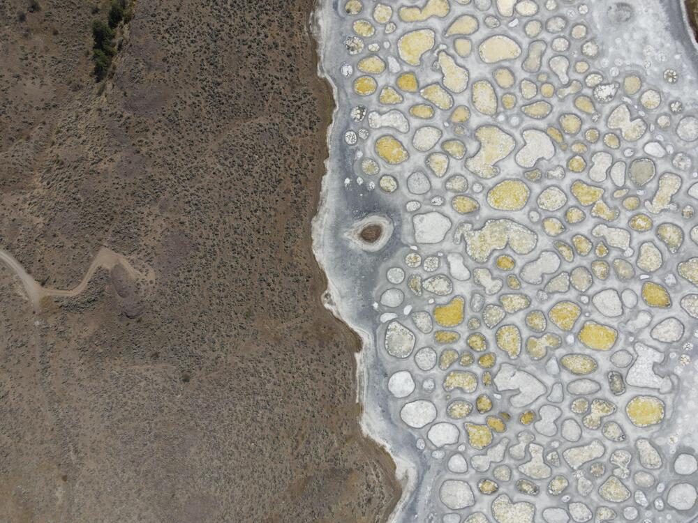 Aerial view of a landscape with dry brown terrain on the left and a salt flat with white and yellow mineral deposits in circular patterns on the right.