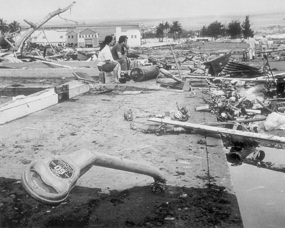 Black and white photo of people sitting amid debris and destruction near water after a disaster; a giant novelty key lies in the foreground amongst scattered wreckage.
