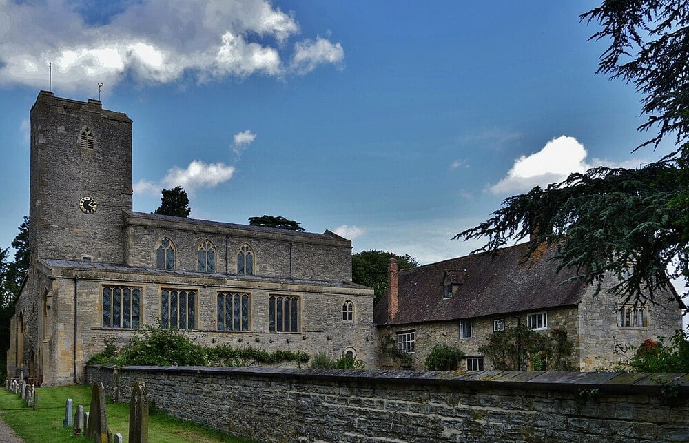 A stone church with a clock tower stands next to an old house, surrounded by a stone wall and a grassy area with gravestones, under a partly cloudy blue sky.
