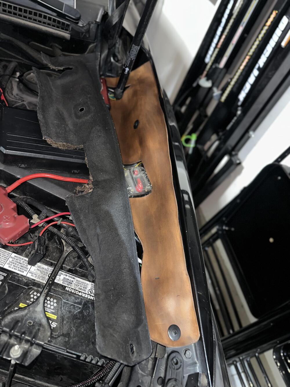 Two engine bay covers, one black and one brown, are placed on a car’s fender near the open hood, with visible red wires and various engine components beneath.