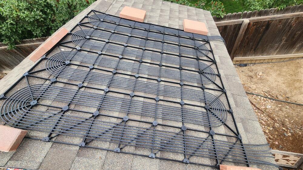 A black plastic solar pool heater mat is secured on a rooftop with bricks at each corner. The mat consists of coiled tubes arranged in a grid pattern to absorb sunlight for heating water.