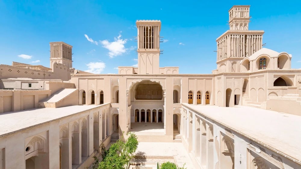 Historic Persian architecture with tall windcatchers, arched doorways, and beige adobe walls under a bright blue sky; a courtyard with greenery is visible below.