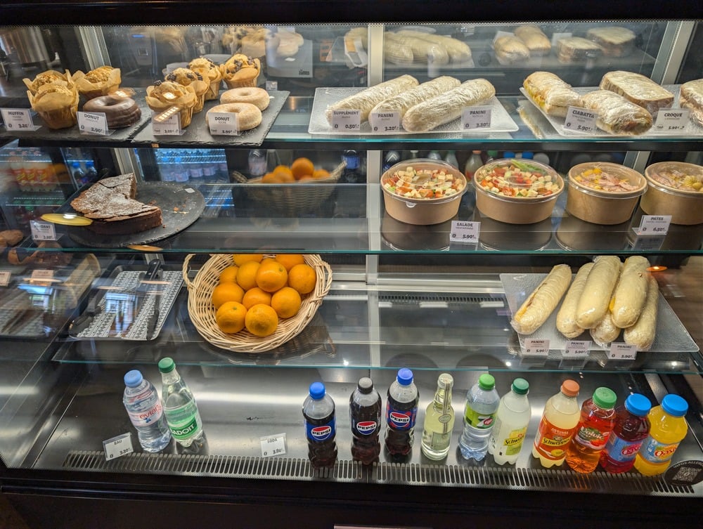 A display case showing sandwiches, pastries, a slice of cake, salads, a basket of oranges, and various bottled drinks including water, soda, and juice.