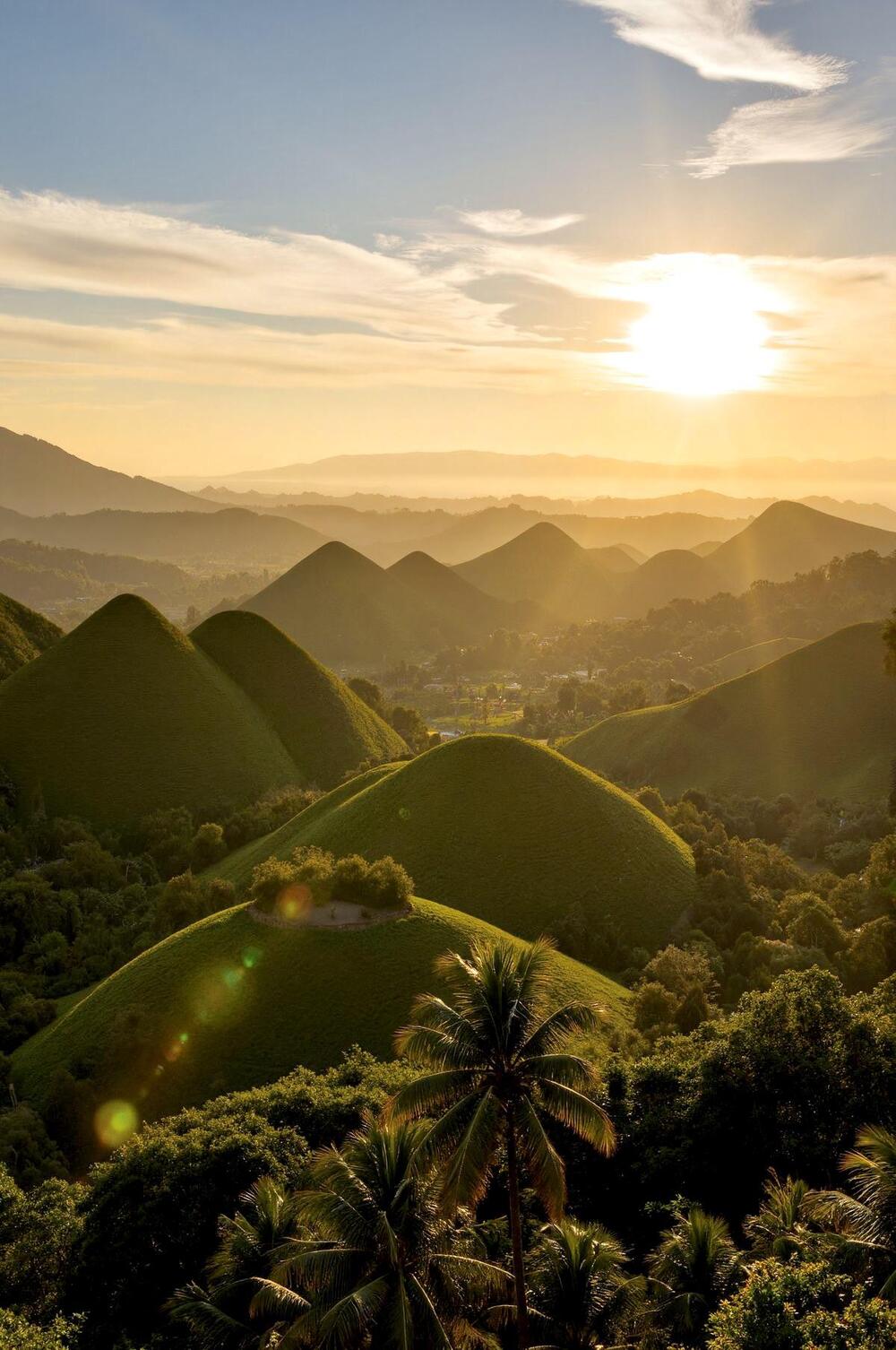Golden sunlight illuminates the lush, rounded hills and valleys of a tropical landscape, with palm trees in the foreground and a hazy mountain range in the distance under a partly cloudy sky.