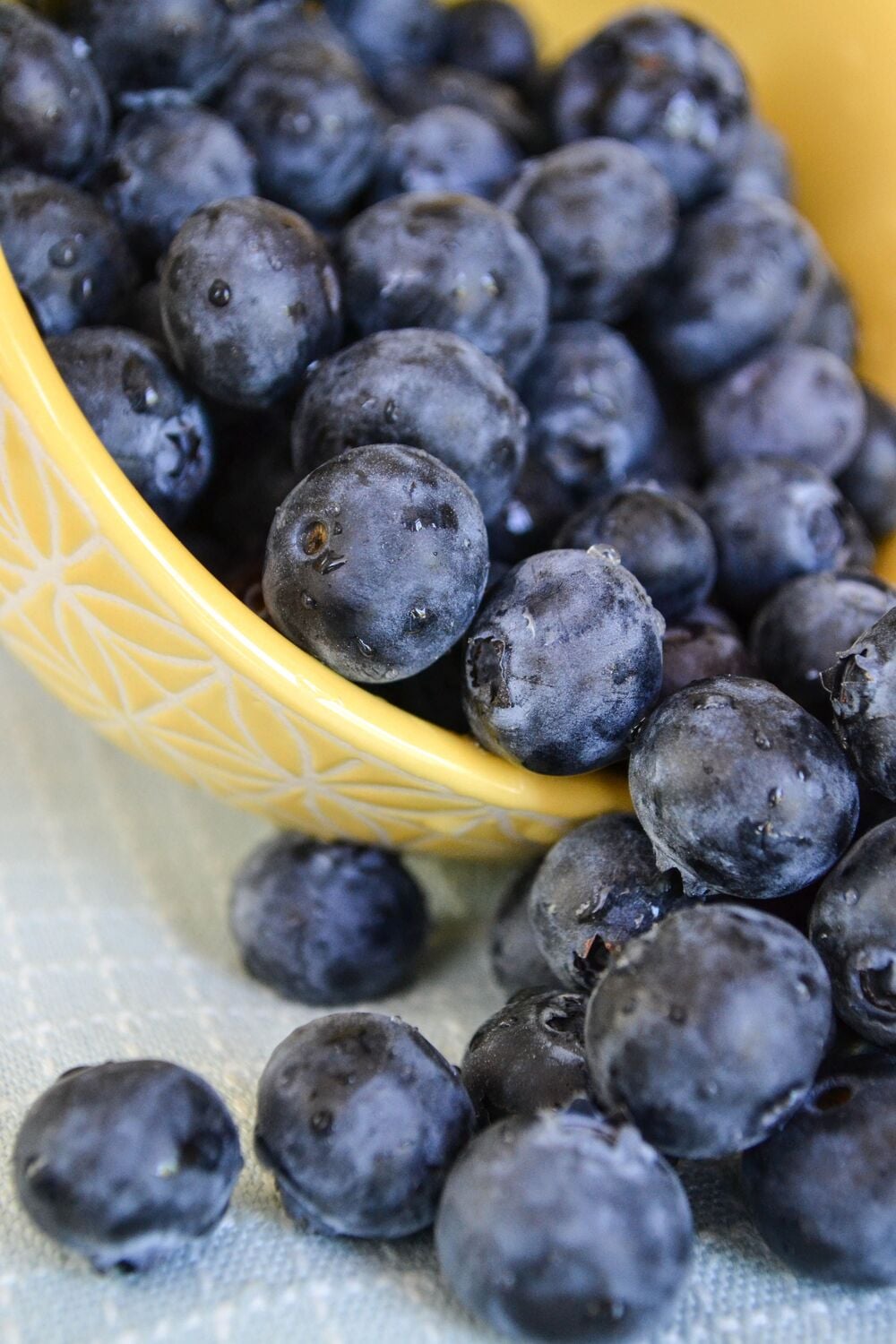A yellow bowl tipped over, spilling fresh blueberries onto a blue and white cloth.