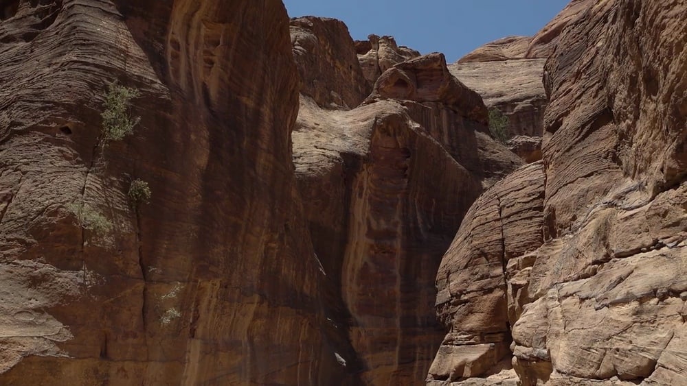 Tall, rugged sandstone cliffs rise under a clear blue sky, with shadows accentuating the layered rock formations and sparse vegetation growing on the steep surfaces.