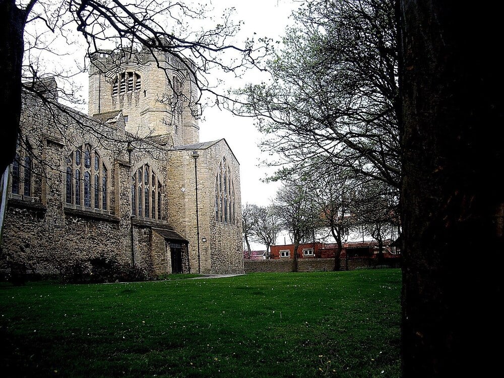 A stone church with tall arched windows stands beside a grassy lawn and leafless trees under an overcast sky.