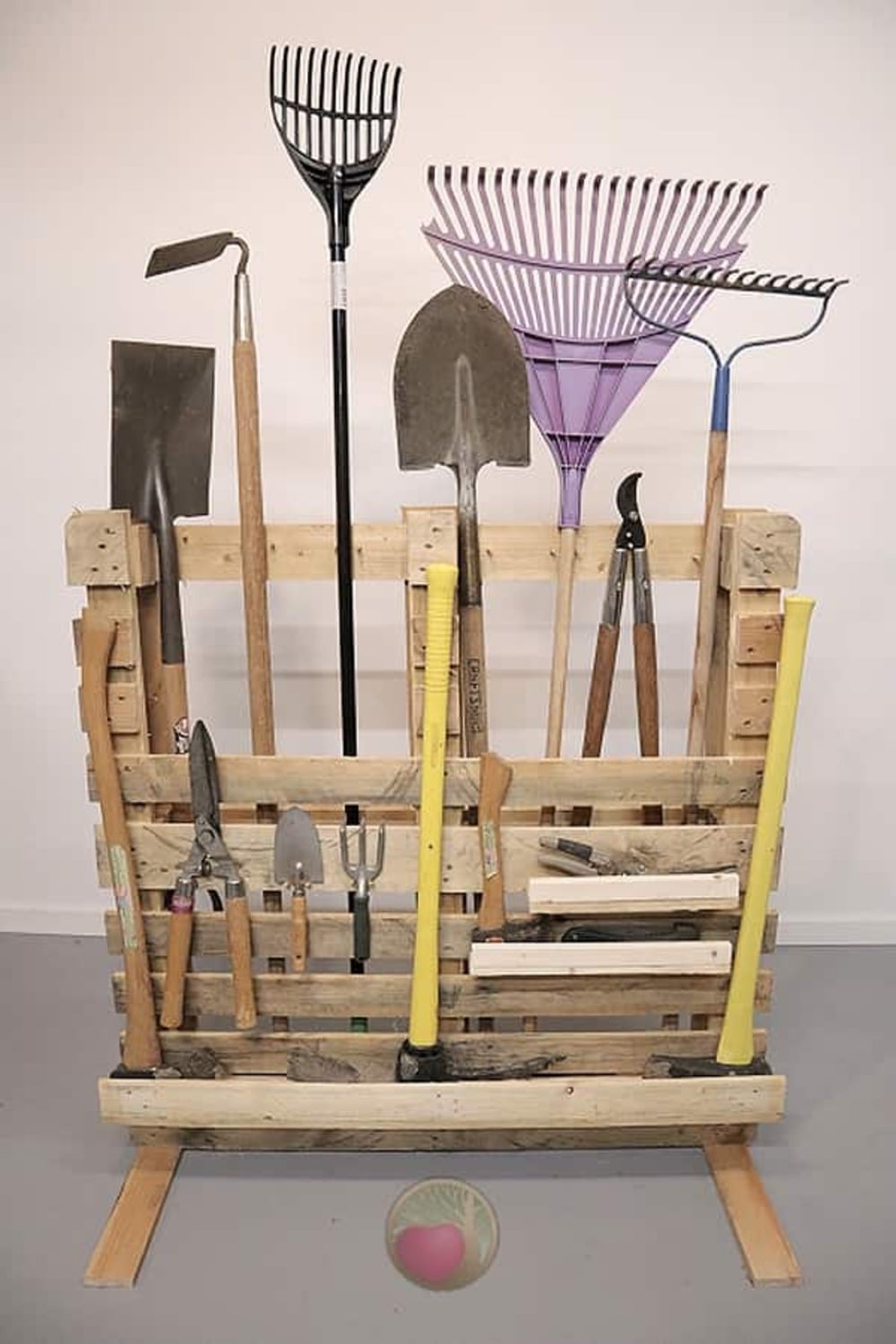 Wooden pallet holding various gardening tools including shovels, rakes, and hand tools, all organized vertically. A pink and green ball is placed in front of the pallet on the floor.
