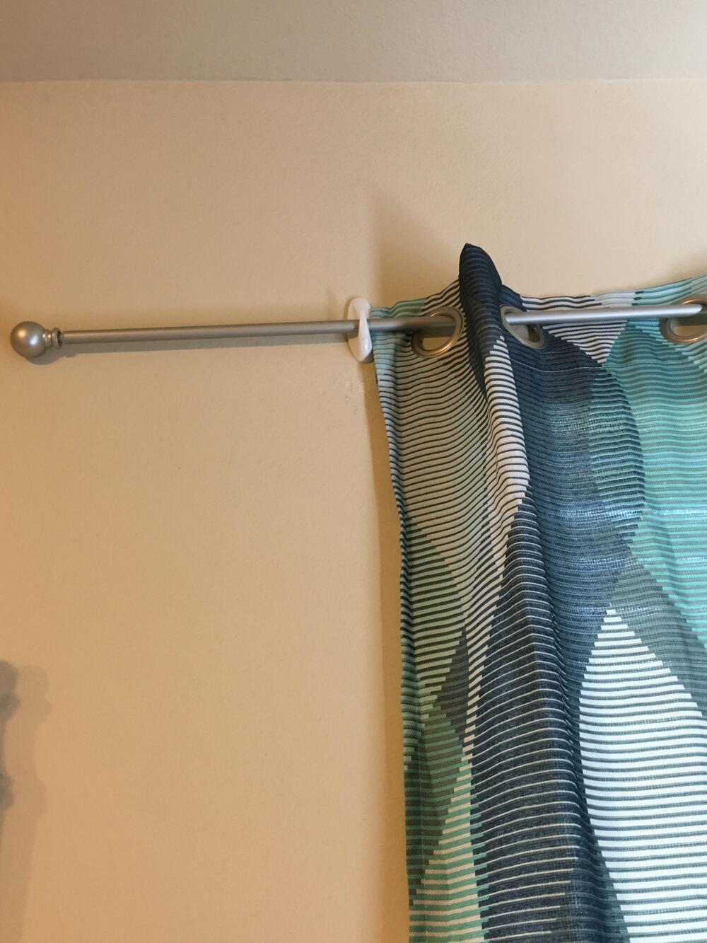 A silver curtain rod is loosely supported by a white zip tie attached to a beige wall, with blue and white patterned curtains hanging from it.