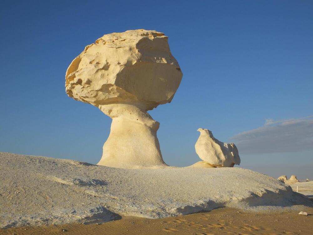 Large, mushroom-shaped white rock formation stands beside a smaller rock in a sandy desert under a clear blue sky.