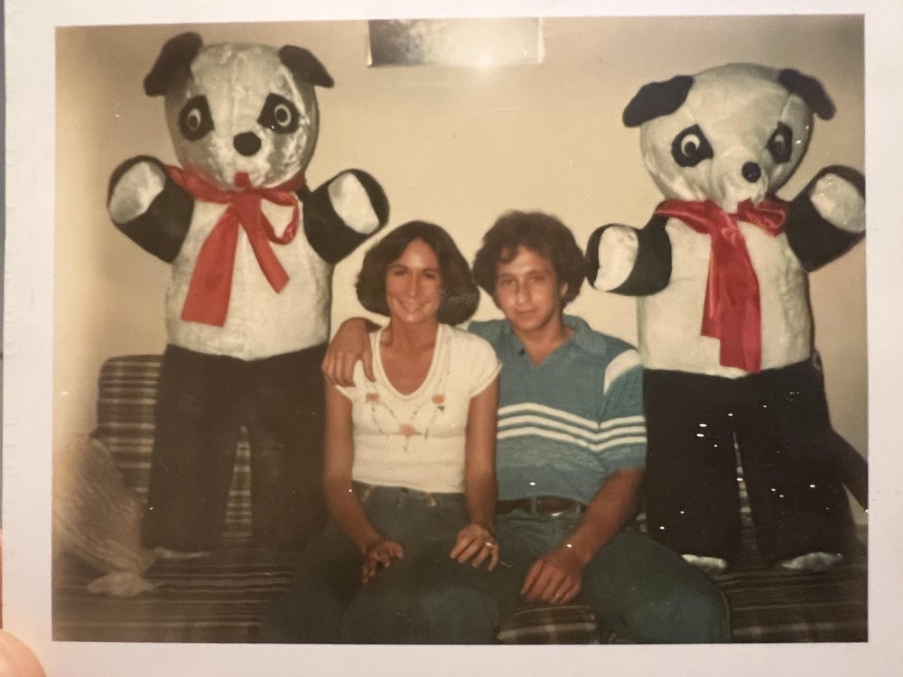 Two people sit on a couch with their arms around each other, flanked by two large stuffed panda bears with red bows, posing for a photo in what appears to be a living room.