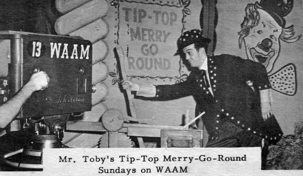 A man in a star-patterned costume gestures beside a large TV camera, with a sign reading “Tip-Top Merry Go Round” and a clown face in the background. Caption: “Mr. Toby’s Tip-Top Merry-Go-Round Sundays on WAAM.”